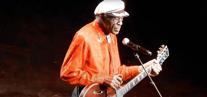 Na foto, Chuck Berry no palco, em destaque. Com uma boina branca e blusa laranja.