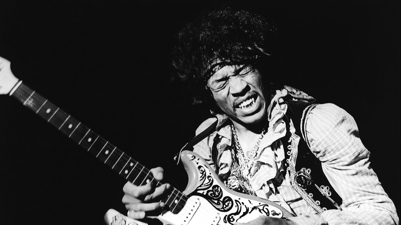 Jimi Hendrix, tocando guitarra com os olhos fechados. A foto é em preto e branco.