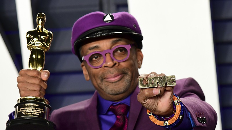 Na foto, Spike Lee, com um terno e boina roxo, além do óculos. Ele está segurando um Oscar e olhando para o lado.