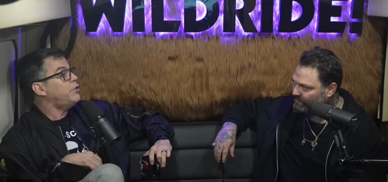 steve-o à direita, bam margera à esquerda, sentados em um sofá preto e a parede do fundo é de pelo cor de caramelo.